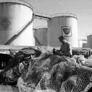 4th July 1975:  A fisherman attending to nets beside oil storage containers belonging to BP on the coast at Aberdeen
