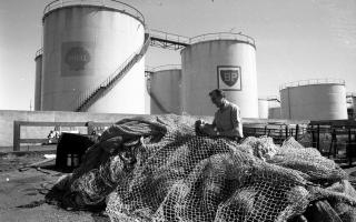 4th July 1975:  A fisherman attending to nets beside oil storage containers belonging to BP on the coast at Aberdeen